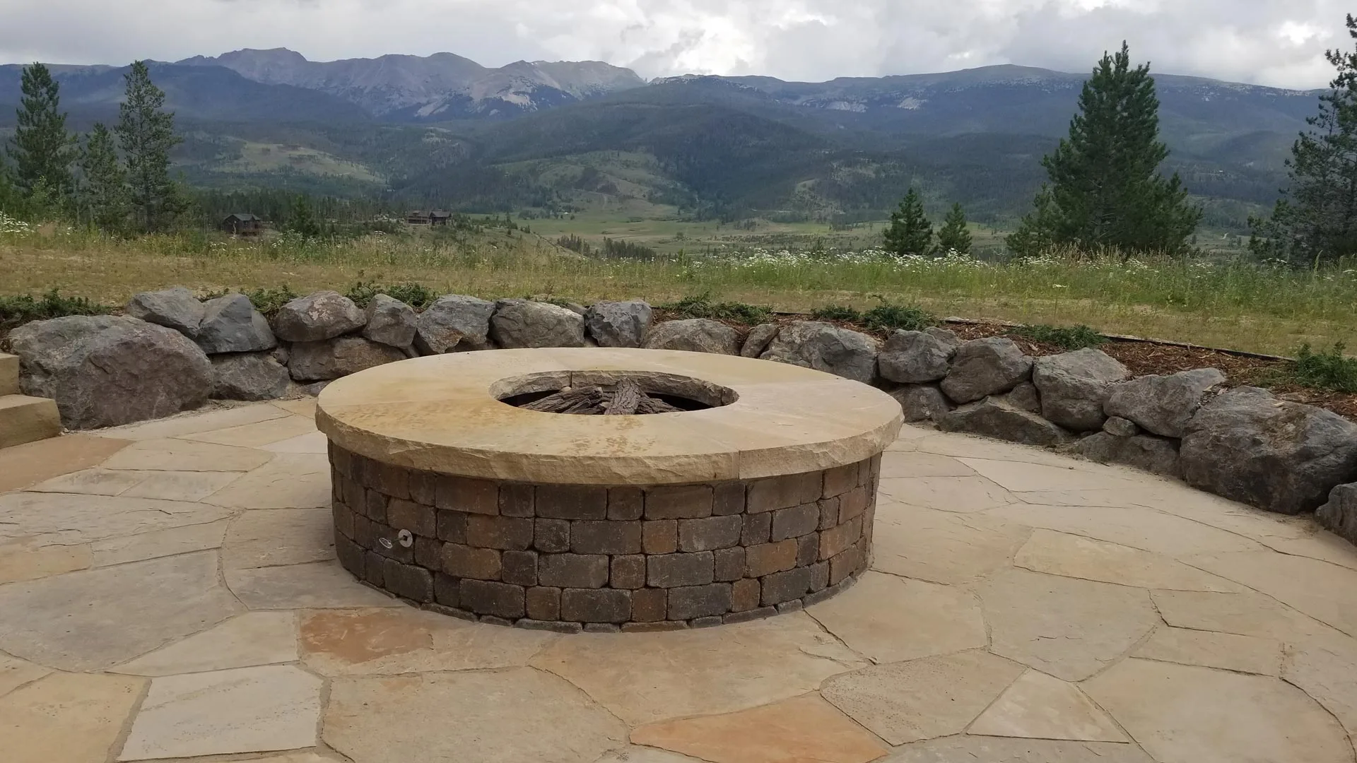 A round fire pit with a flagstone patio and rock retaining wall overlooking the mountains in Winter Park, CO.